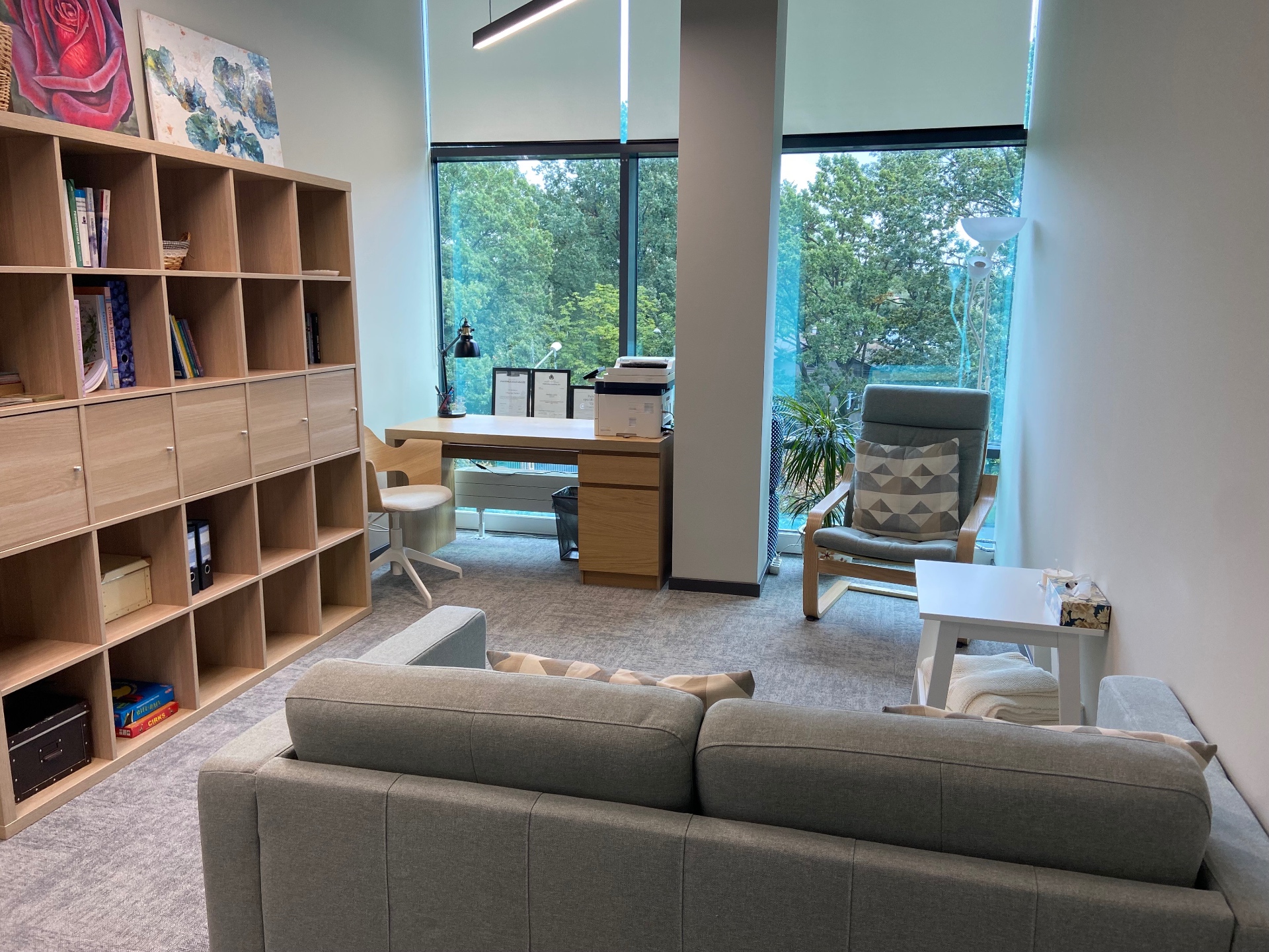 Viktorijas Zariņas kabinets Rīgā, Vienības gatvē 109 — gaiša telpa ar dīvānu, plauktiem un lielu logu uz kokiem